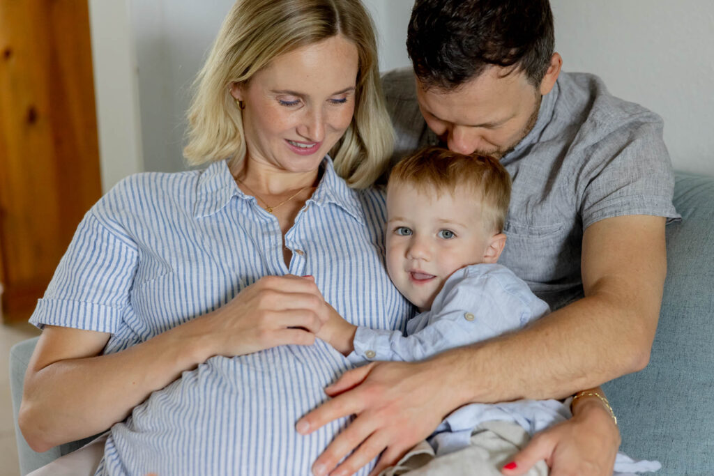 Eine blonde schwangere Frau und ihr Mann kuscheln mit ihrem Sohn.